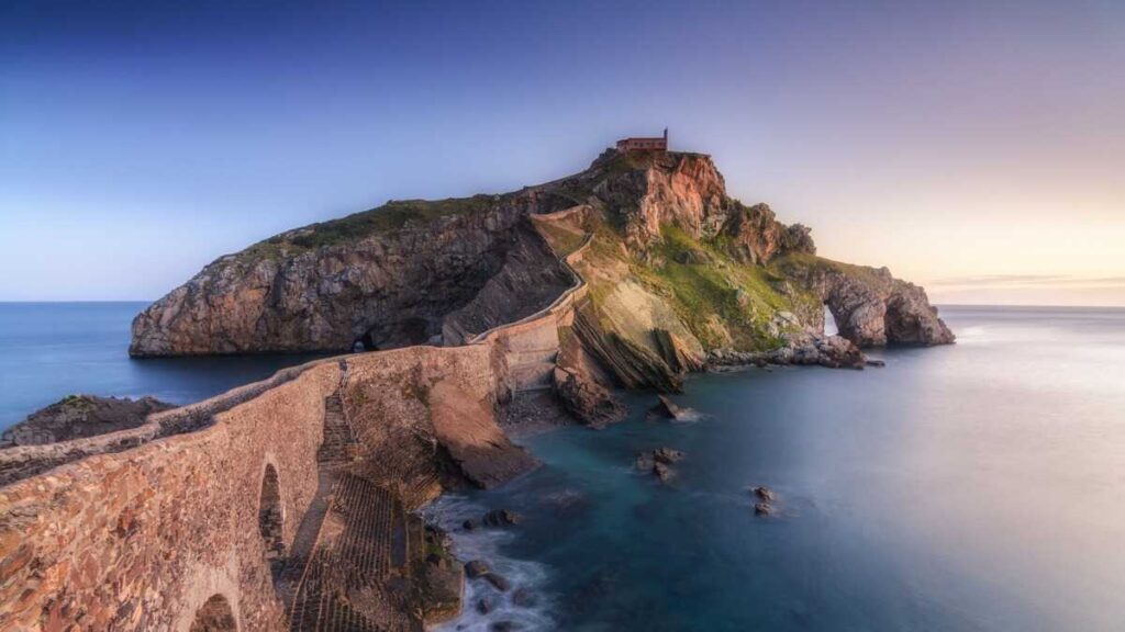 The San Juan de Gaztelugatxe stone bridge connecting to a small island with a purple sky sunset reflecting on the water