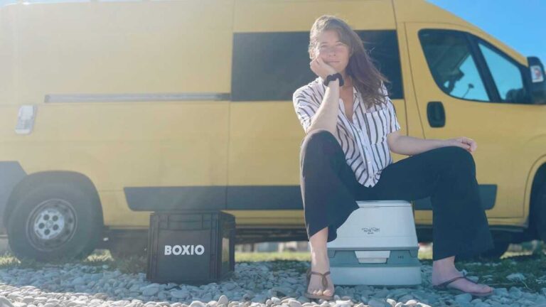 Lady with long brown hair and blue eyes, sitting on a chemical toilet with a Boxio dry toilet beside and a yellow van in the background