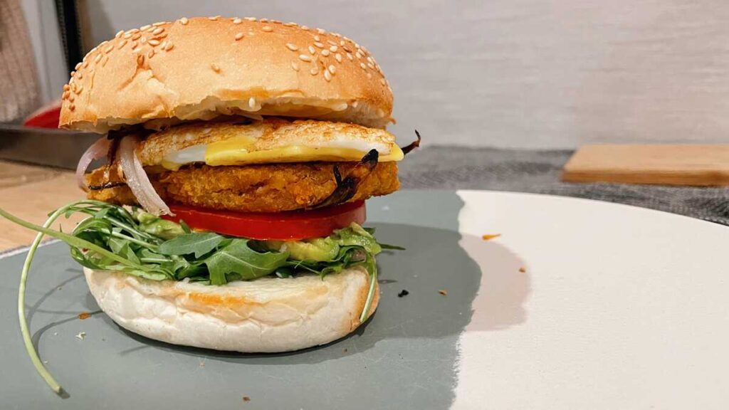 burger made from a sesame seed brioche bun, lettuce, tomato, vegetarian kroket burger, egg, and onion, on a blu and white bamboo plate