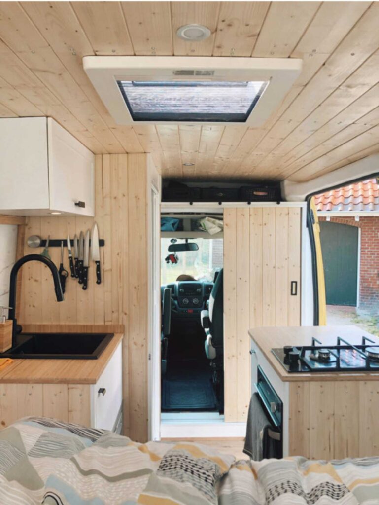 Campervan interior with wooden roof and walls, white cabinetry, and black accessories