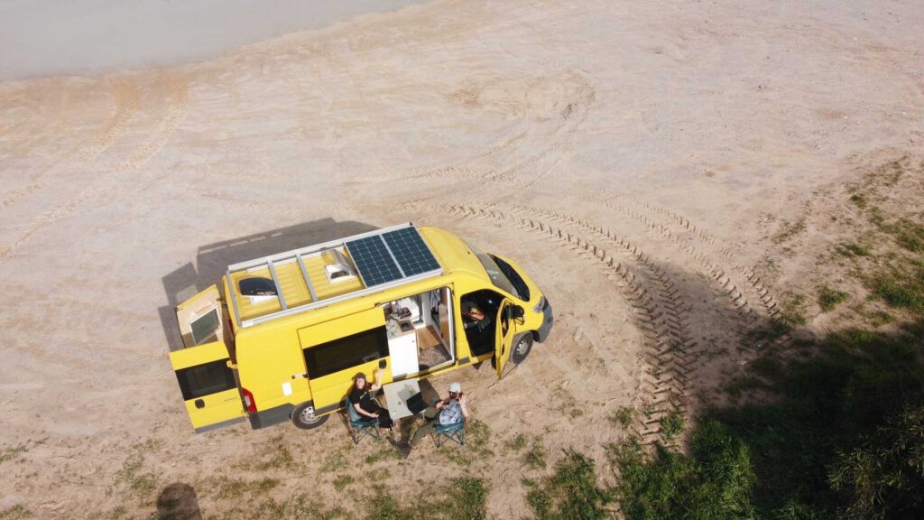 Drone image of a yellow van parked on the sand with 2 people and a picnic set