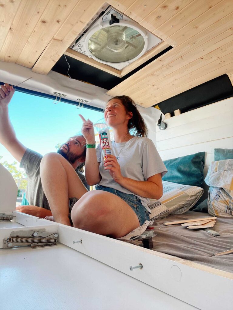 Two people sitting inside a DIY campervan inspecting the tongue and groove roof panels with a glue pistol in hand