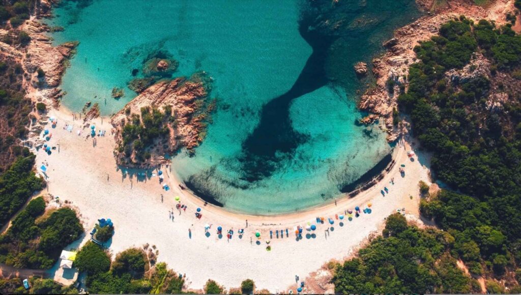 Aerial view of a crescent-shaped beach in Sardinia with turquoise water and surrounding rocky terrain.