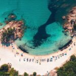 Aerial view of a crescent-shaped beach in Sardinia with turquoise water and surrounding rocky terrain.